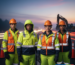 A road crew of four workers wearing compliant hi-vis yellow and orange jackets and vests with reflective tape, standing at a construction site at dusk with long traffic light trails in the background.